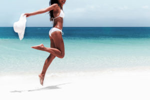 Beach Ready Bikini Body Sexy Slim Legs And Toned Thighs And Butt. Suntan Happy Woman Jumping In Freedom On White Sand With Sun Hat. Weight Loss Success Or Epilation Concept.